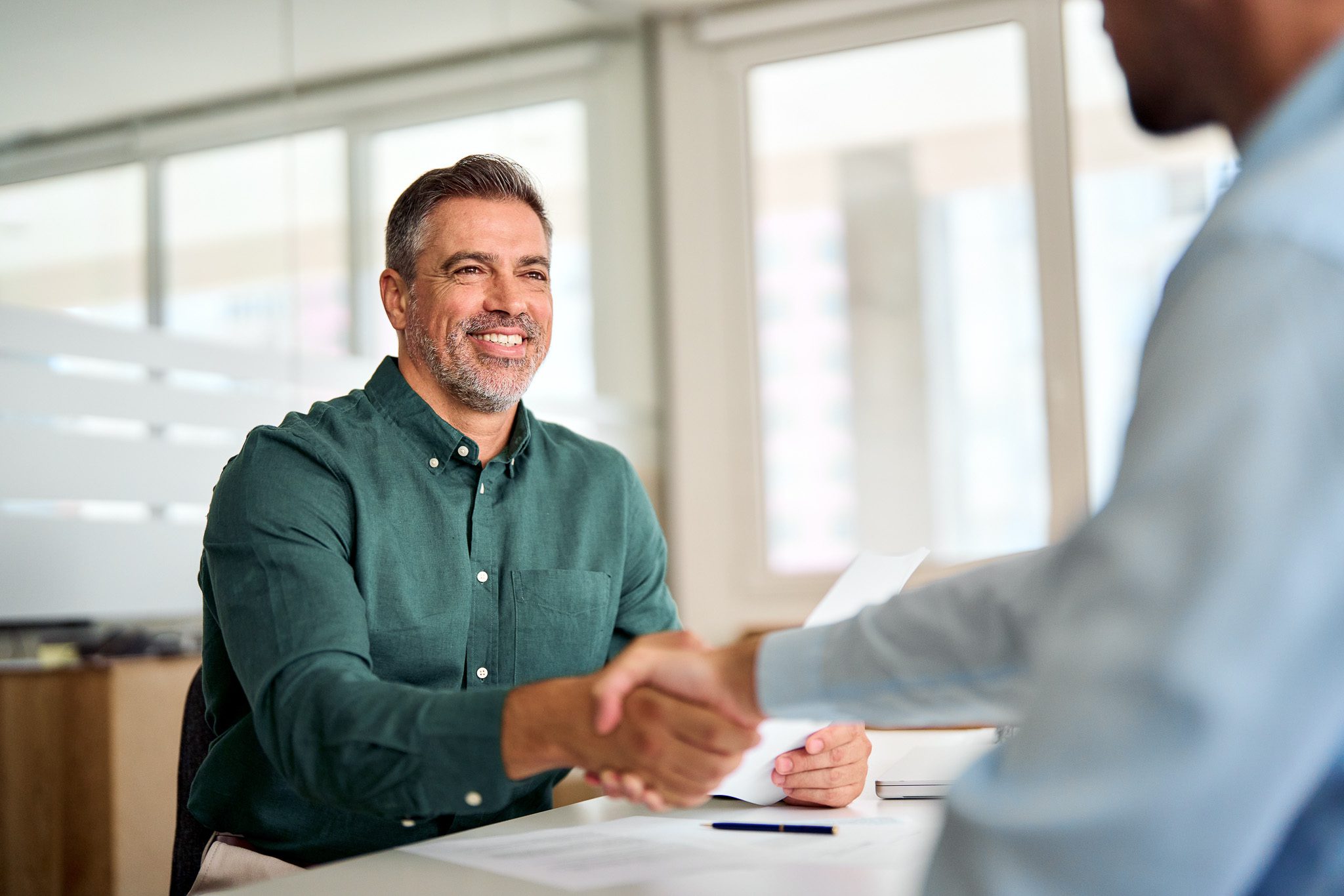 A man in a green shirt smiles while shaking hands with another person in an office setting.