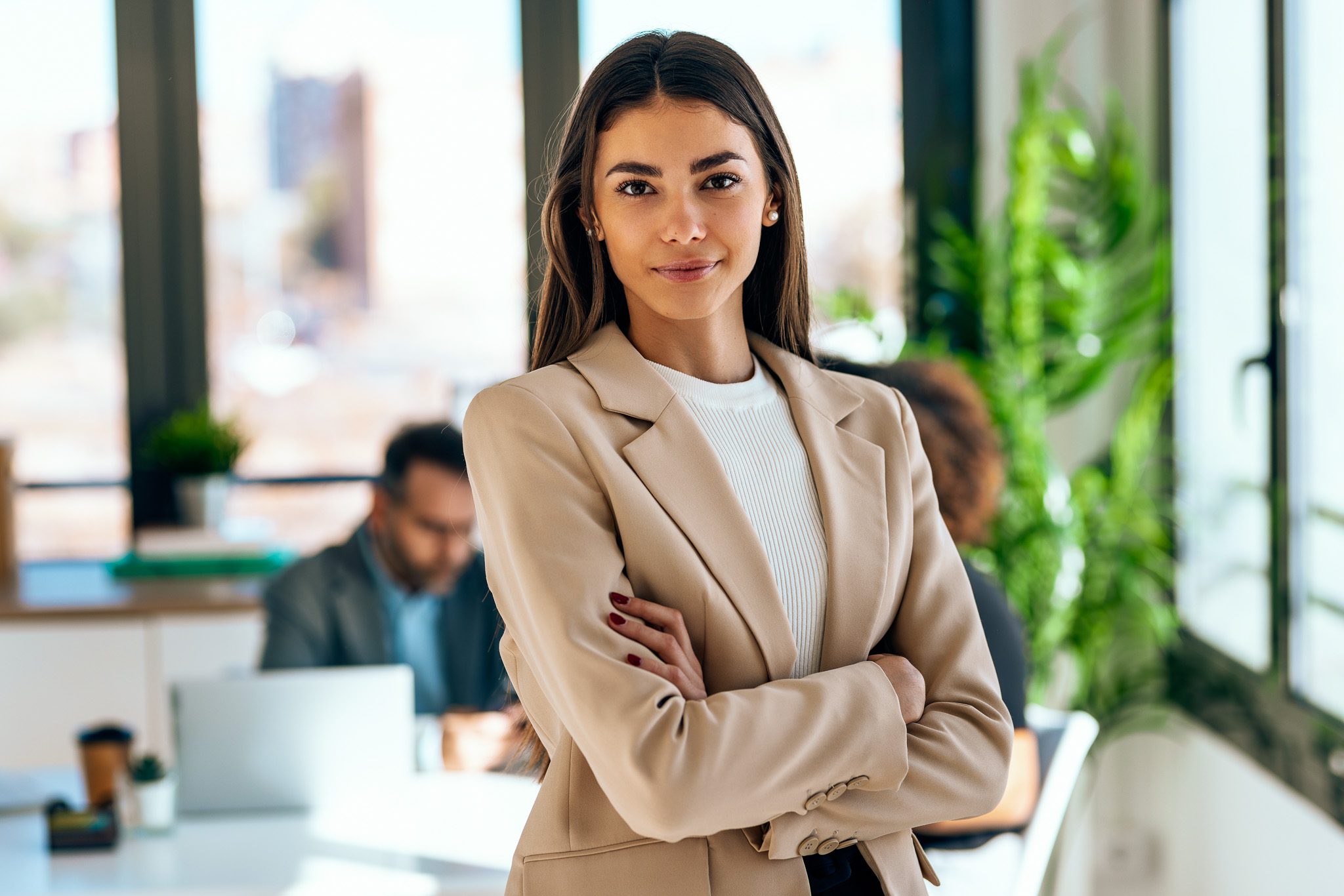 A woman in a beige blazer stands confidently with arms crossed in a bright office. Two people work on laptops in the background.